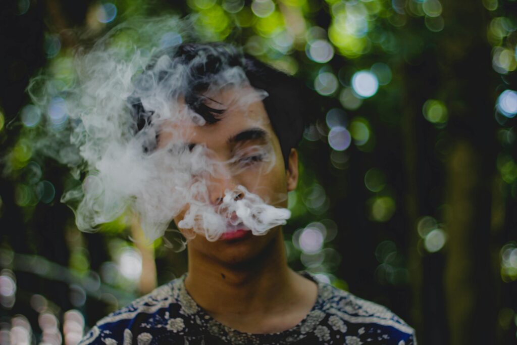 A young man smoking and exhaling smoke outdoors with a blurred natural background.