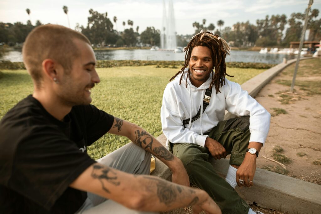 Two men sitting and smiling together by the lake, enjoying a sunny day outdoors.